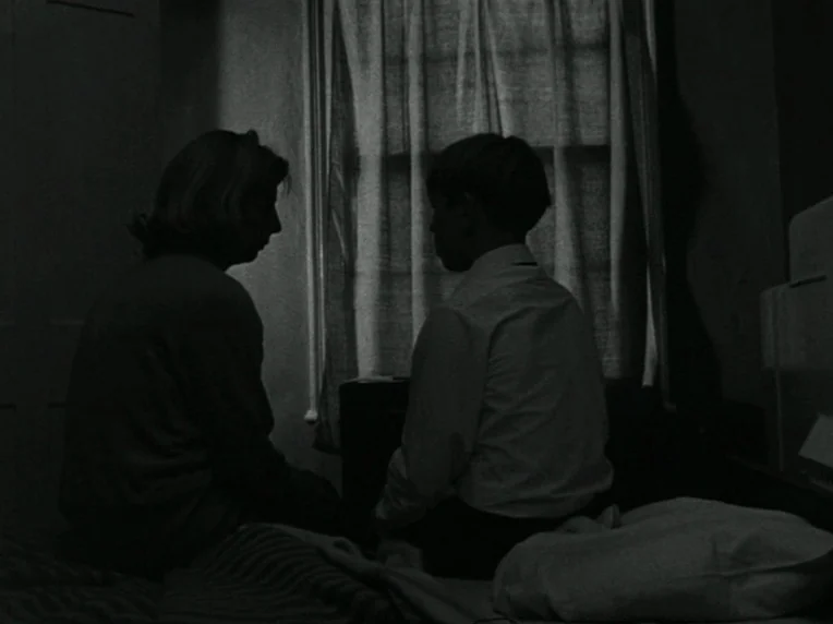 Children (1976) movie still: medium — A dark, sparsely furnished room features a window covered by thin curtains. A woman and a child sit on a bed,…
