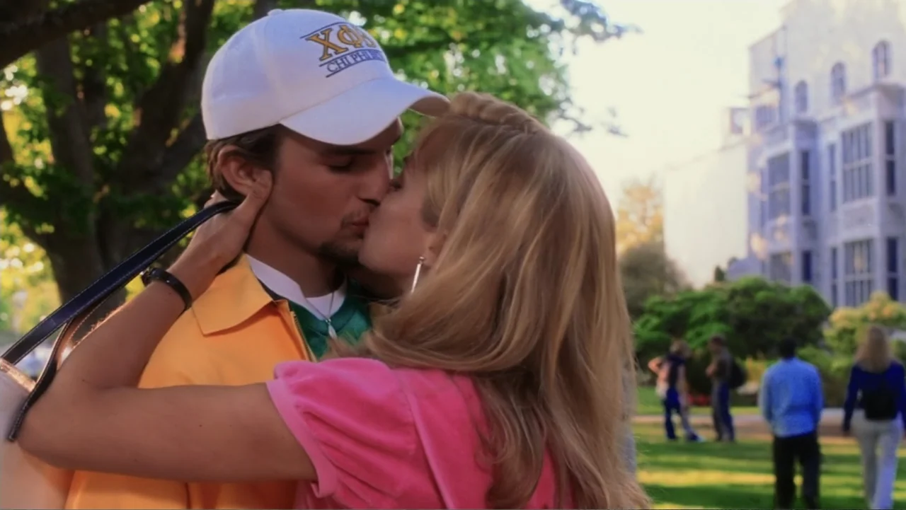 The Butterfly Effect (2004) movie still: medium — A sunny college campus path is lined with large trees and an academic building. A young man and a…