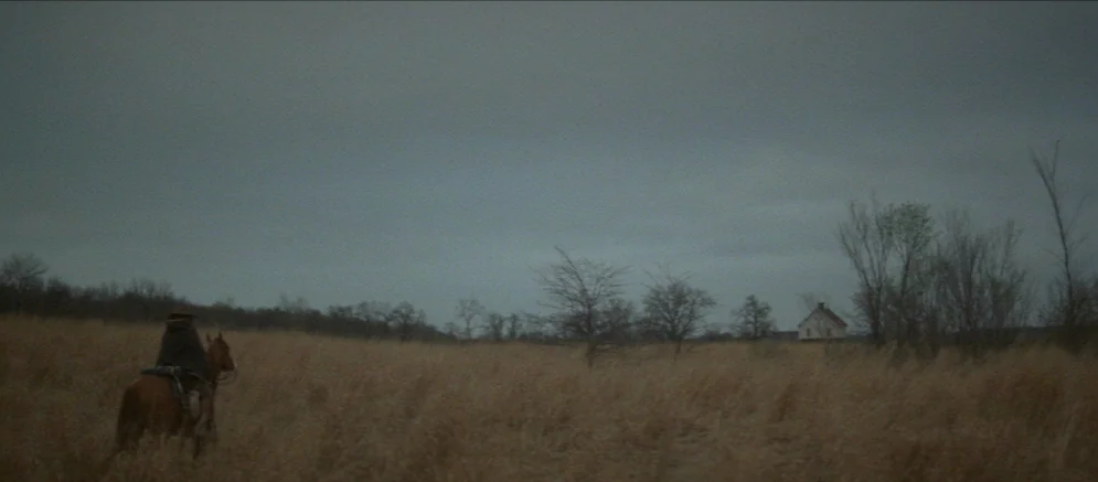 Ride With The Devil movie still: wide — A vast, open field of dry grass sits under a dense, overcast gray sky. A lone figure on horseback moves…