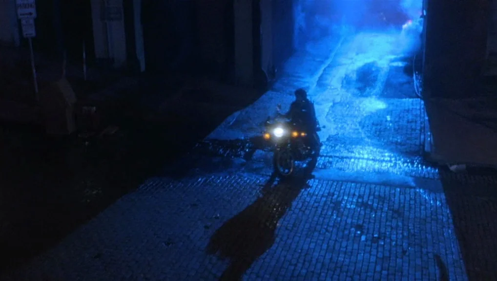 Graffiti Bridge (1990) movie still: wide — A dark, brick-paved urban path filled with blue smoke. A motorcyclist approaches the camera.
