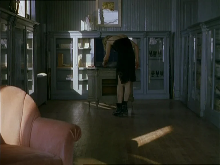 Last Days (2005) movie still: wide — A room with wooden floors and tall gray glass-fronted cabinets lining the walls. A woman in a black dress leans…
