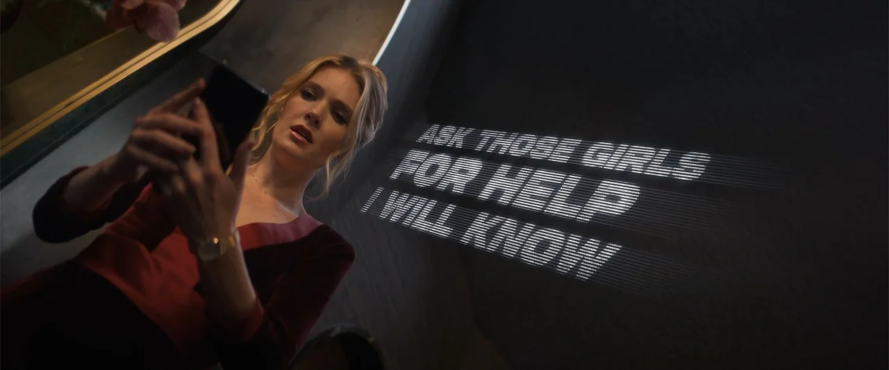 Drop (2025) movie still: medium — A dark, dimly lit corridor features text projected directly onto a plain grey wall. A woman stands in the…