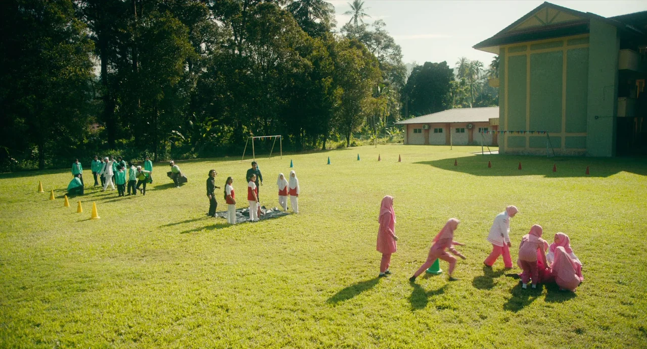 Tiger Stripes (2023) movie still: wide — A sunny school playing field with green grass sits adjacent to a large building. Several students in…