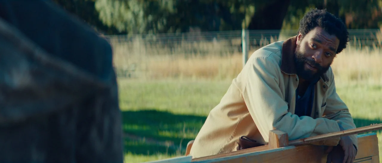 Z for Zachariah (2015) movie still: medium — A man stands behind a wooden fence in a sunny grassy field. He leans on the top rail while looking…