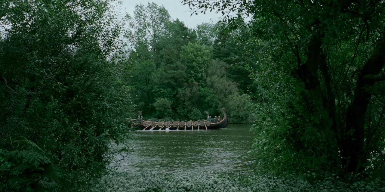 The Northman (2022) movie still: wide — A wooden Viking longship traverses a calm, narrow river surrounded by dense green woodland. The boat is…