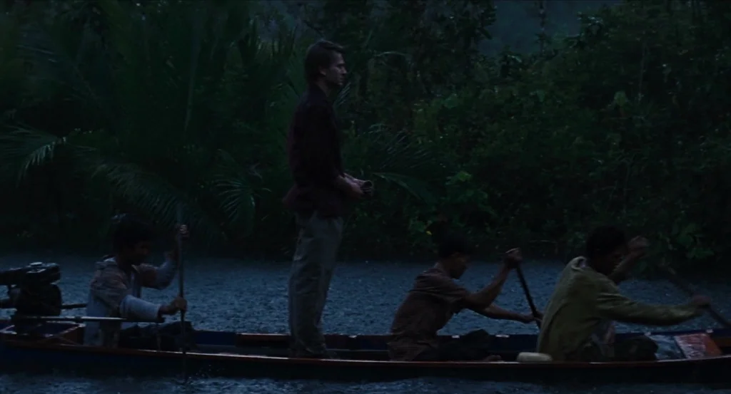 Almayer’s Folly (2011) movie still: wide — A dark, rainy river scene is filled with dense jungle foliage along the banks. Four men in a long, narrow…