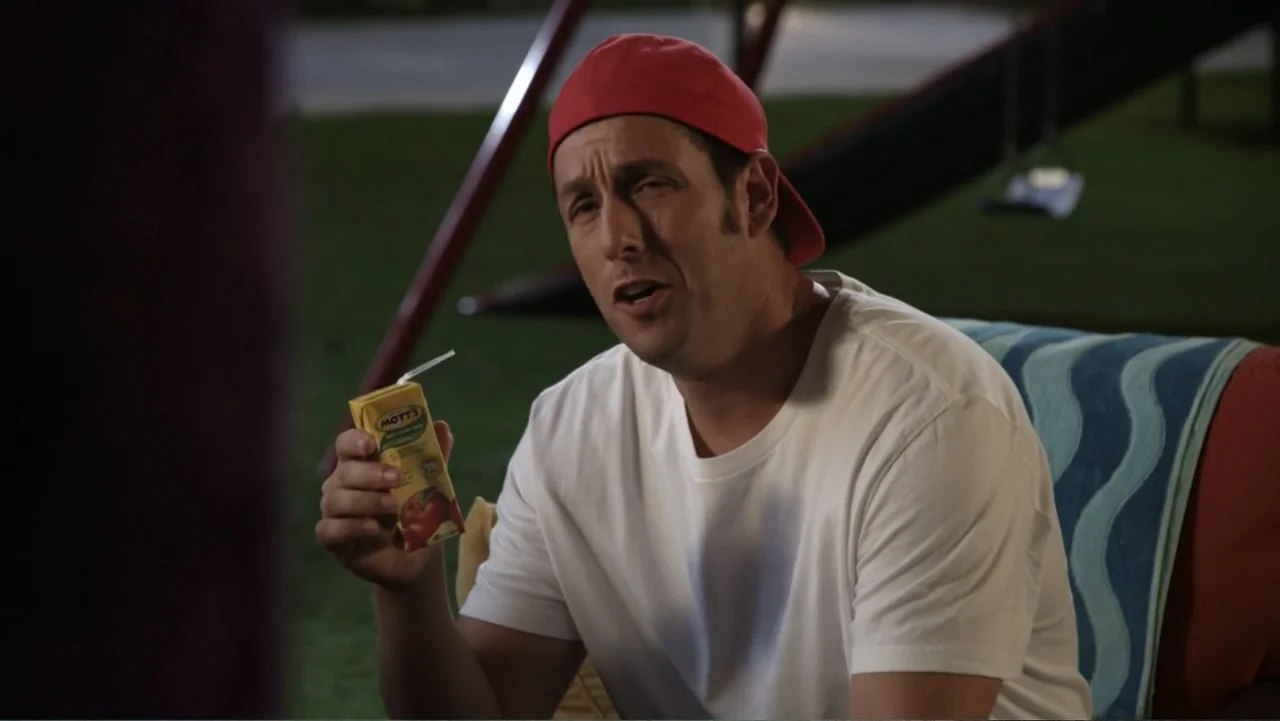 Grown Ups 2 (2013) movie still: medium — A dark playground at night features a man sitting on a towel-covered surface. He holds a yellow juice box…