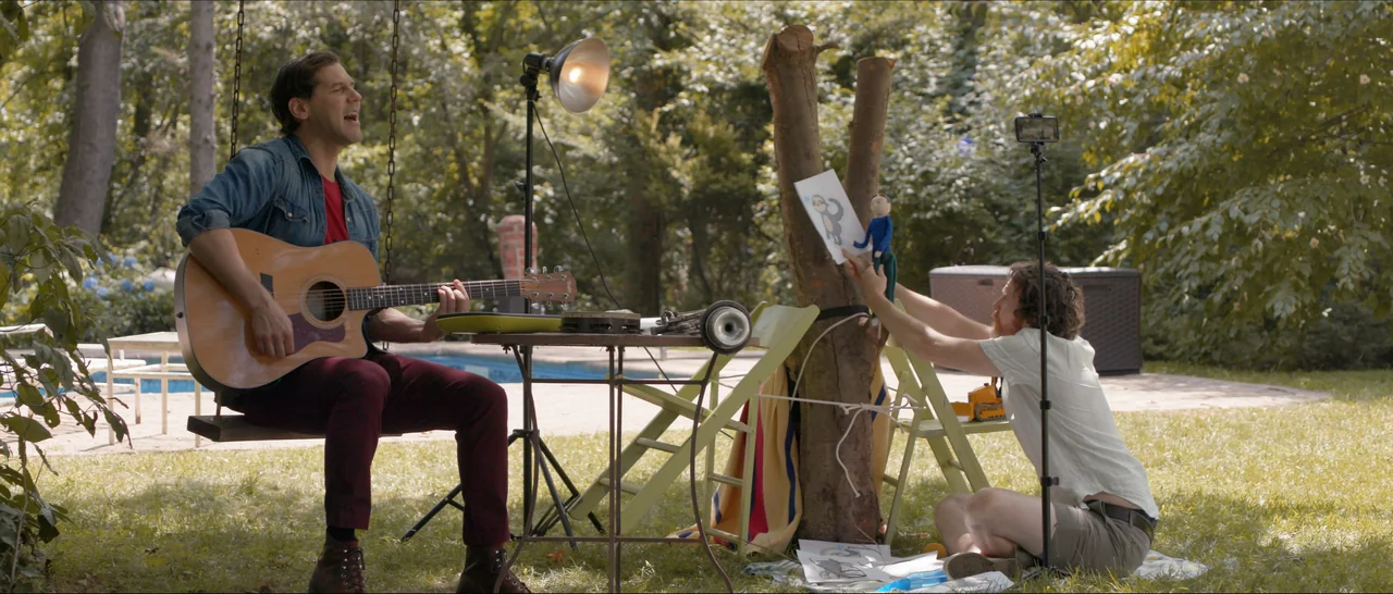 Songs For A Sloth (2021) movie still: wide — A sunny backyard setting features a guitar player on a swing and a man filming. The man filming sits on…