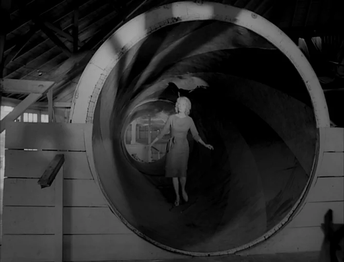 Carnival of Souls (1962) movie still: medium — A woman walks through a large wooden tube located inside a barn-like structure. Sunlight illuminates…