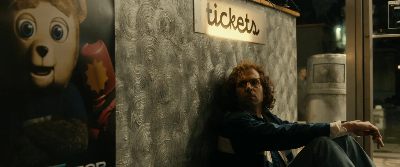 Brigsby Bear (2017) movie still: medium — A man sits on the carpeted floor of a vintage movie theater lobby. The wall behind him features a circular…