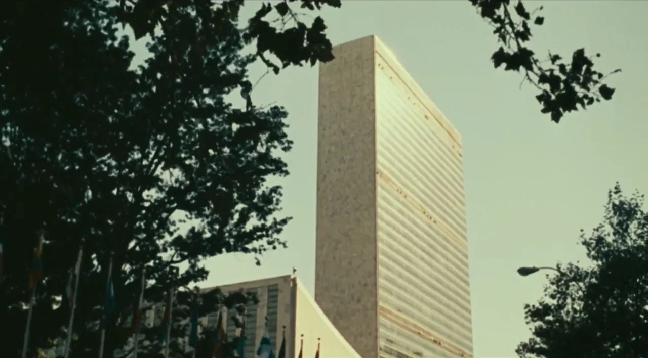 The Other Conquest (1999) movie still: establishing — The United Nations Secretariat Building stands tall against a clear, pale sky. Several flags…