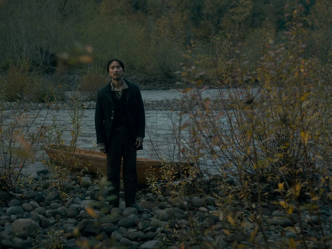 First Cow (2019) movie still: fullbody — A rocky riverbank features a slow-moving river and sparse, autumn-colored shrubs and trees. A man in…