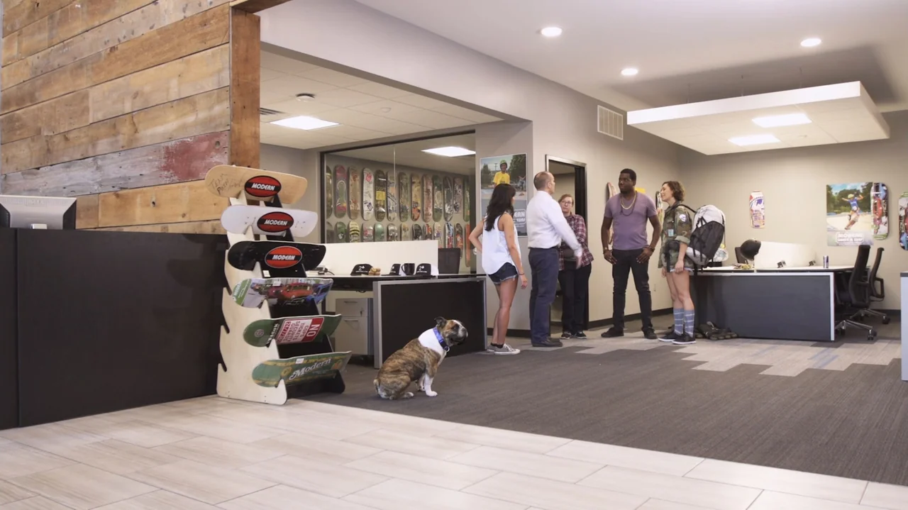 Sk8 Dawg (2018) movie still: wide — An open office area features a wood-paneled wall and a row of skateboards displayed. A group of four adults…