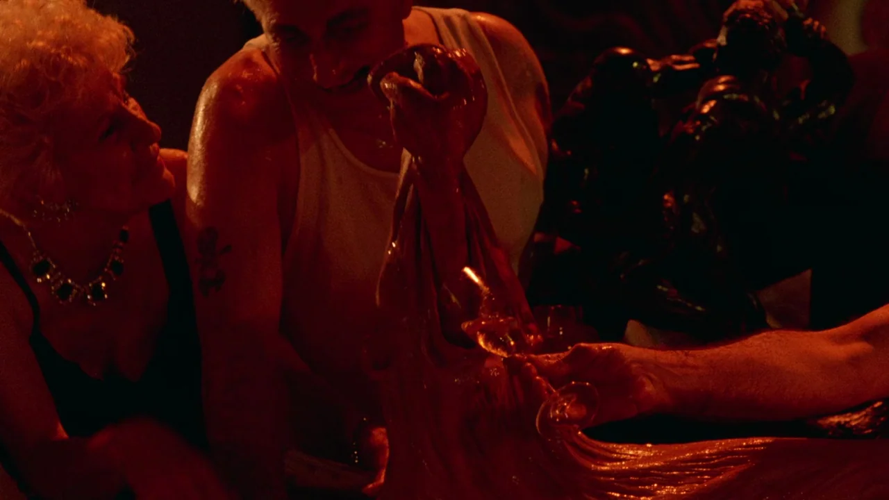 Society (1989) movie still: medium — A dimly lit room with people gathered around a large red mass of food. The man and woman reach their hands into…
