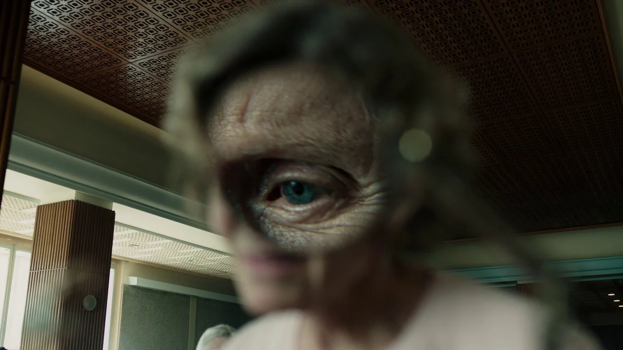 A Cure for Wellness (2017) movie still: closeup — An interior office space with a textured ceiling and a prominent square support pillar. An elderly…