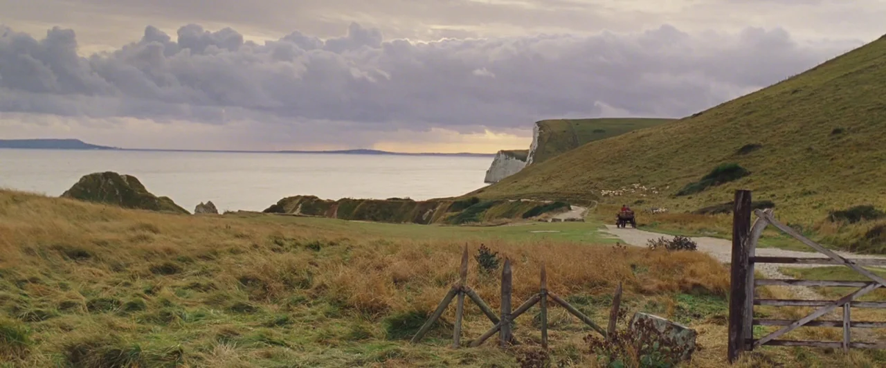 Far from the Madding Crowd (2015) movie still: wide — A grassy coastal cliff overlooks a wide bay under heavy, overcast cloud cover. A lone…