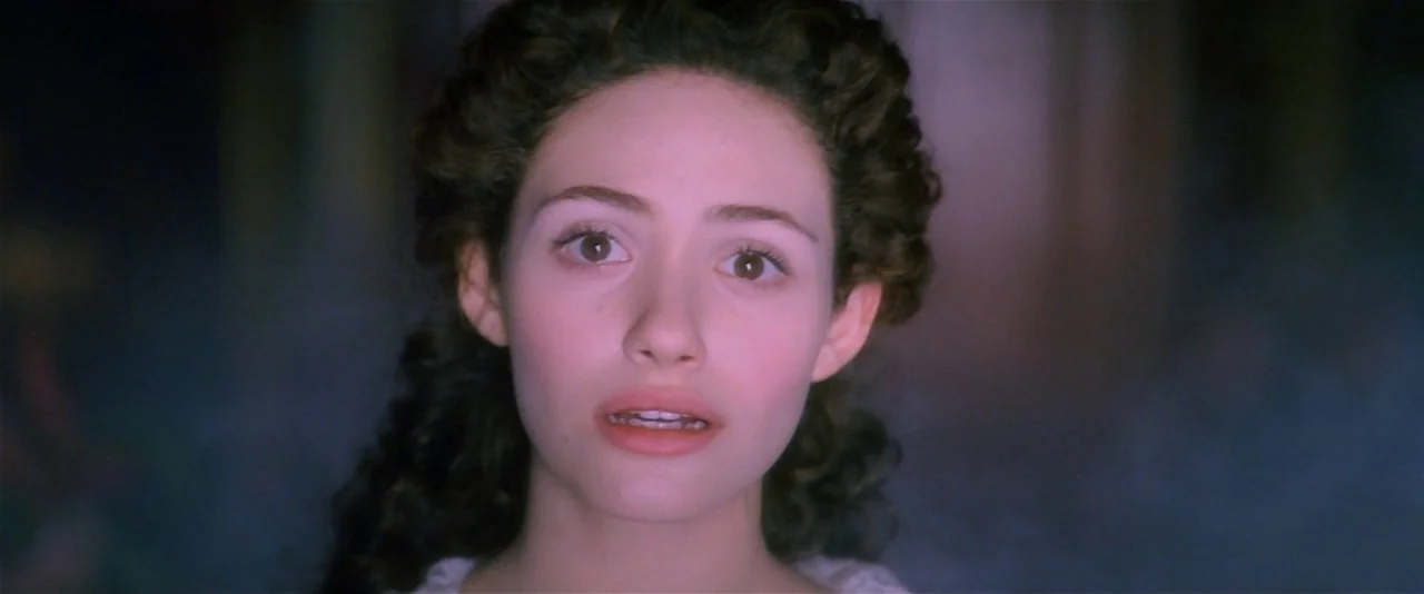 The Phantom of the Opera (2004) movie still: closeup — A dark, misty space surrounds a young woman with dark curly hair. The woman stares forward…
