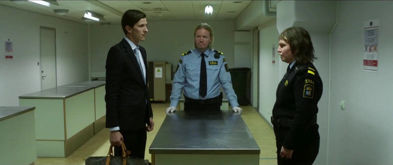 Border movie still: fullbody — An institutional inspection room features light green walls and a central stainless steel table. A man in a suit and…