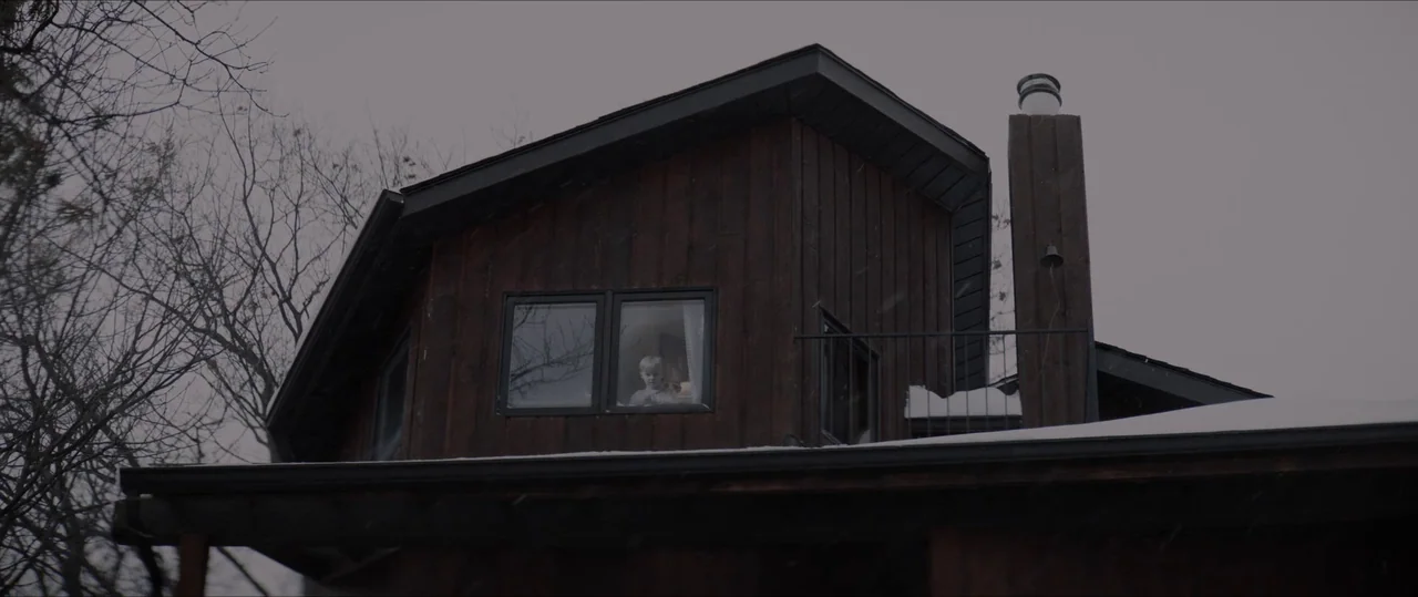 Anything For Jackson (2020) movie still: wide — A dark wooden house with a metal chimney and snowy roof sits under grey skies. A small child watches…