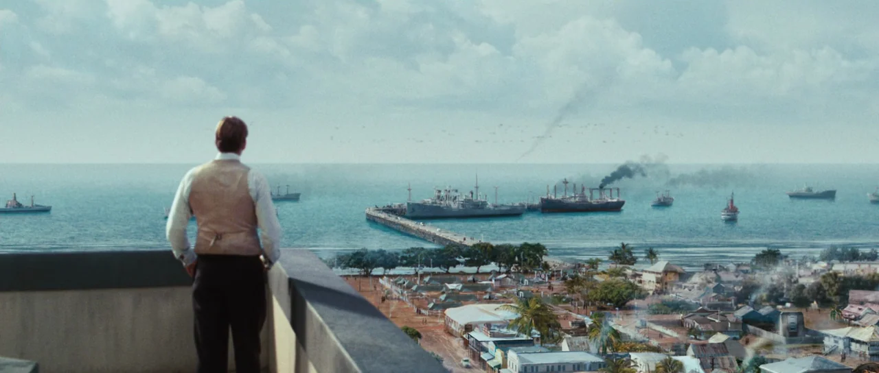 Australia (2008) movie still: wide — A high vantage point overlooks a tropical harbor filled with several large naval ships. A man stands on the…