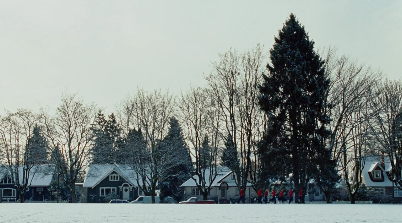Juno (2007) movie still: wide — A snowy suburban landscape with rows of houses behind a large, flat open field. A small group of joggers in bright…
