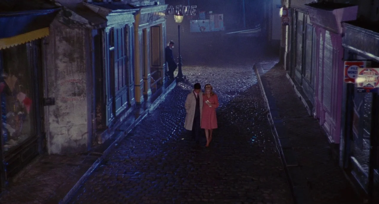 The Umbrellas Of Cherbourg (1964) movie still: wide — A wet, narrow cobblestone street is lined with shopfronts under a dark night sky. A man in a…
