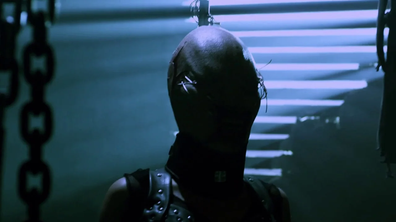 Hellraiser: Revelations movie still: closeup — A dark industrial space features horizontal fluorescent light slats visible in the background. A…