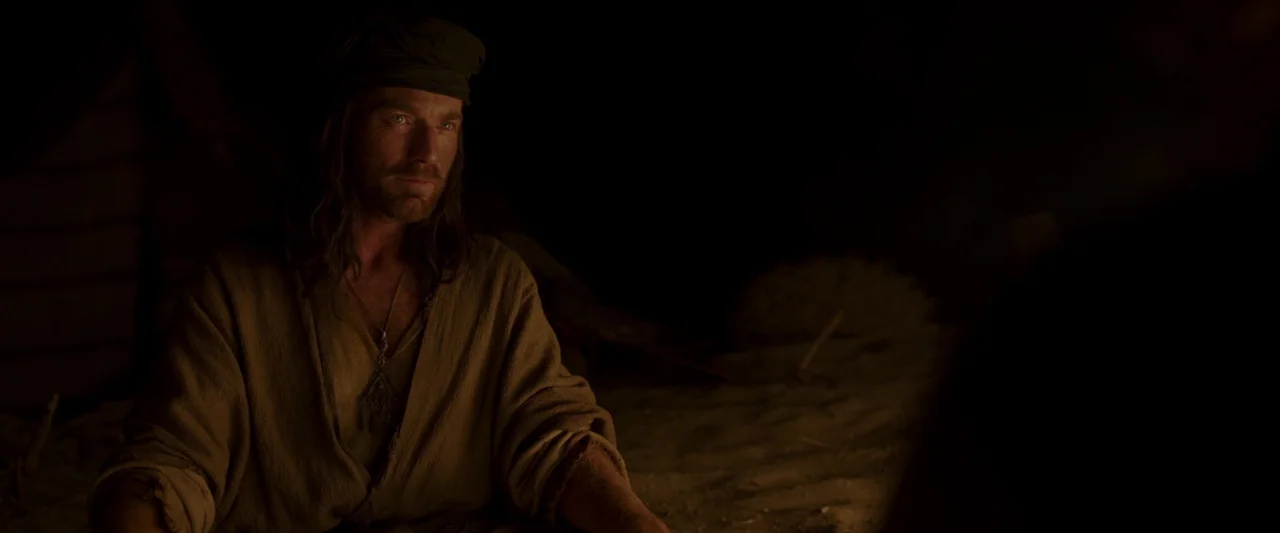 Last Days in the Desert (2015) movie still: medium — A dark night setting features a man seated on sand by an unseen fire. The subject wears…