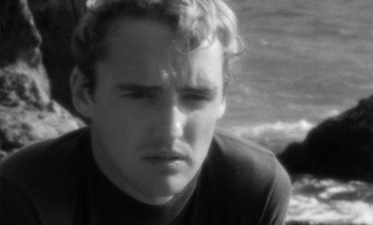 Night Tide (1961) movie still: closeup — A man stands near rocky cliffs beside a crashing ocean. He looks into the distance while the sea waves blur…