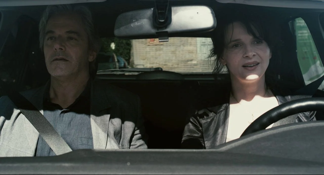 Certified Copy (2010) movie still: medium — A man and a woman ride in the front seats of a vehicle. The woman drives while the man sits in the…