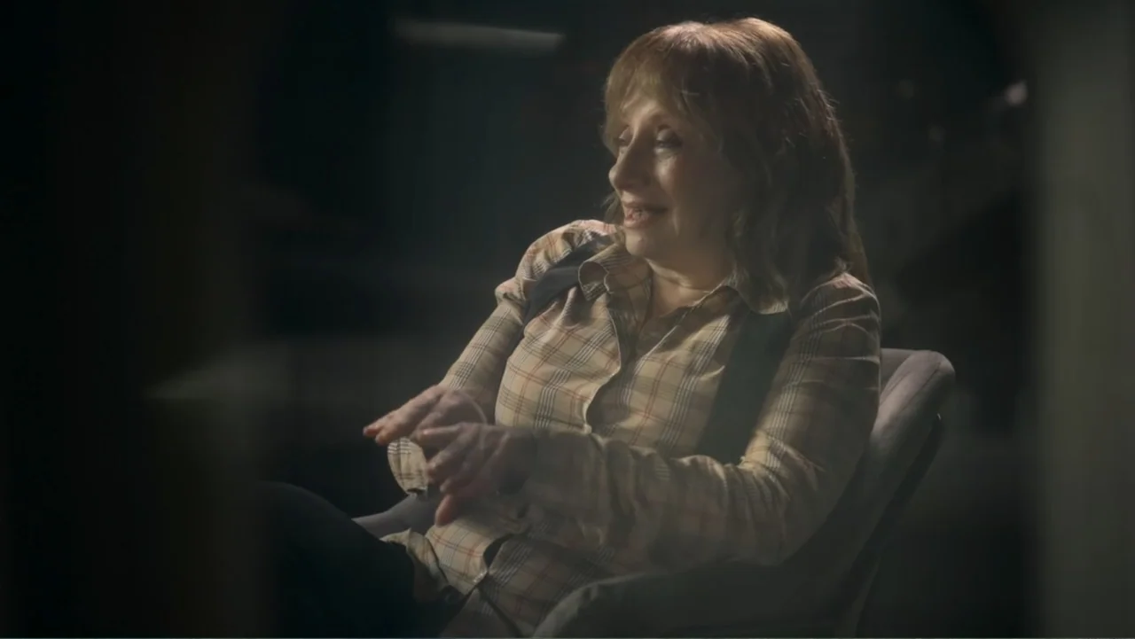 Transfers (2024) movie still: medium — A dimly lit room with a single chair and dark background. A woman in a plaid shirt sits in the chair talking…