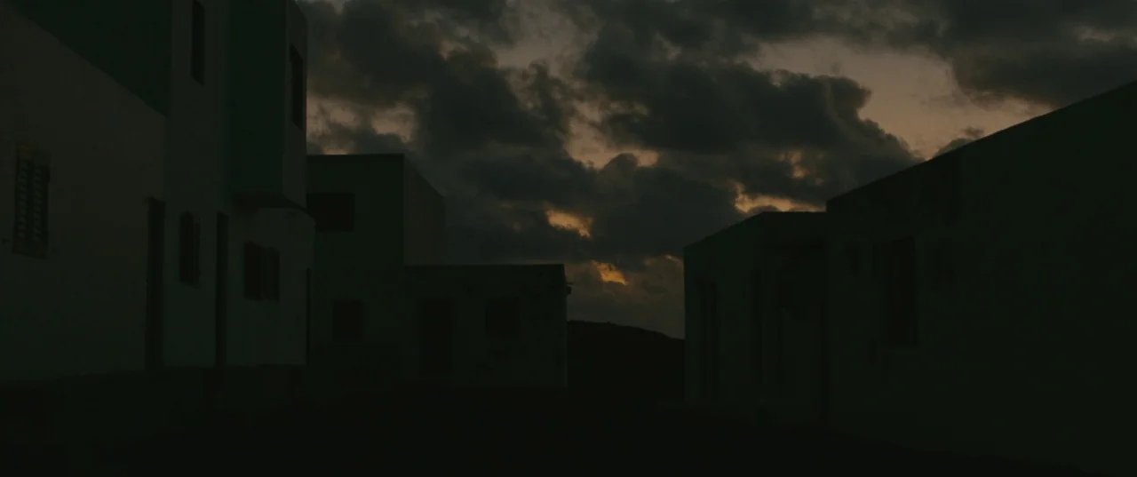 Evolution (2015) movie still: wide — A group of simple, low-rise buildings stands in a dark, rural exterior setting. Thick clouds fill the sky…