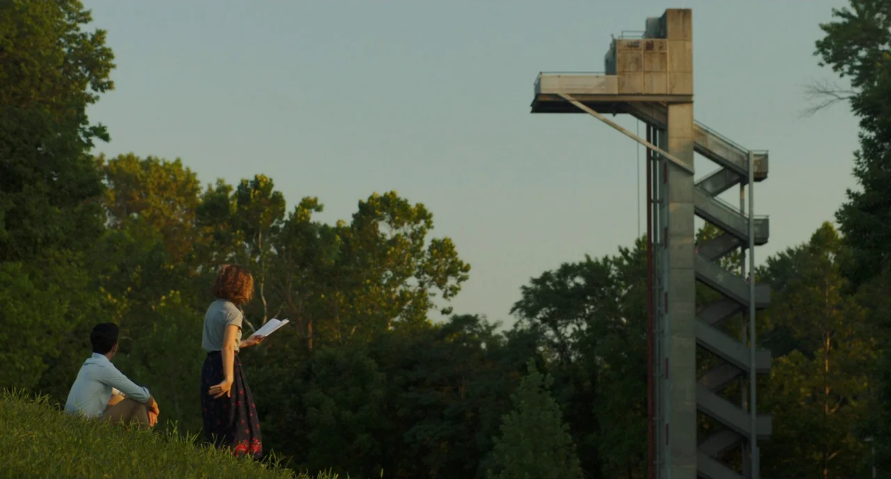 Columbus (2017) movie still: wide — A grassy hill overlooks a tall, industrial-looking concrete tower set against a forest line. A woman reads aloud…
