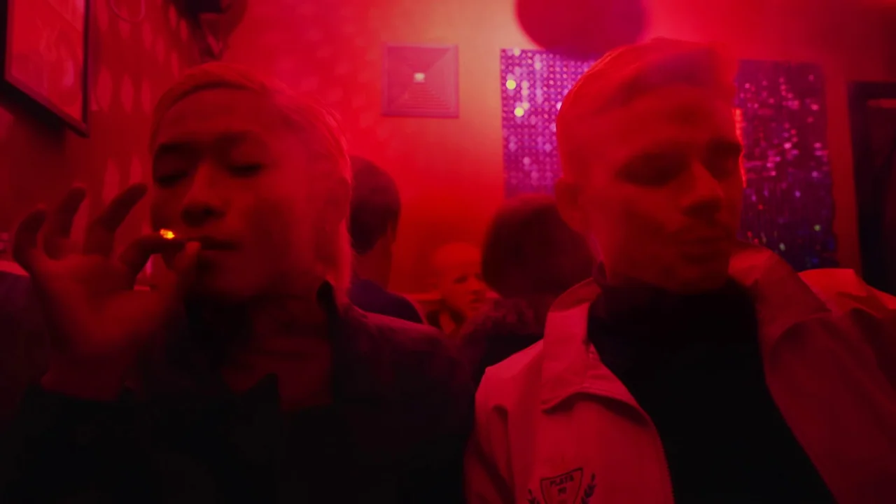 The Last Year of Darkness (2023) movie still: medium — A dimly lit club interior bathed in intense red light with blurred figures behind. Two young…