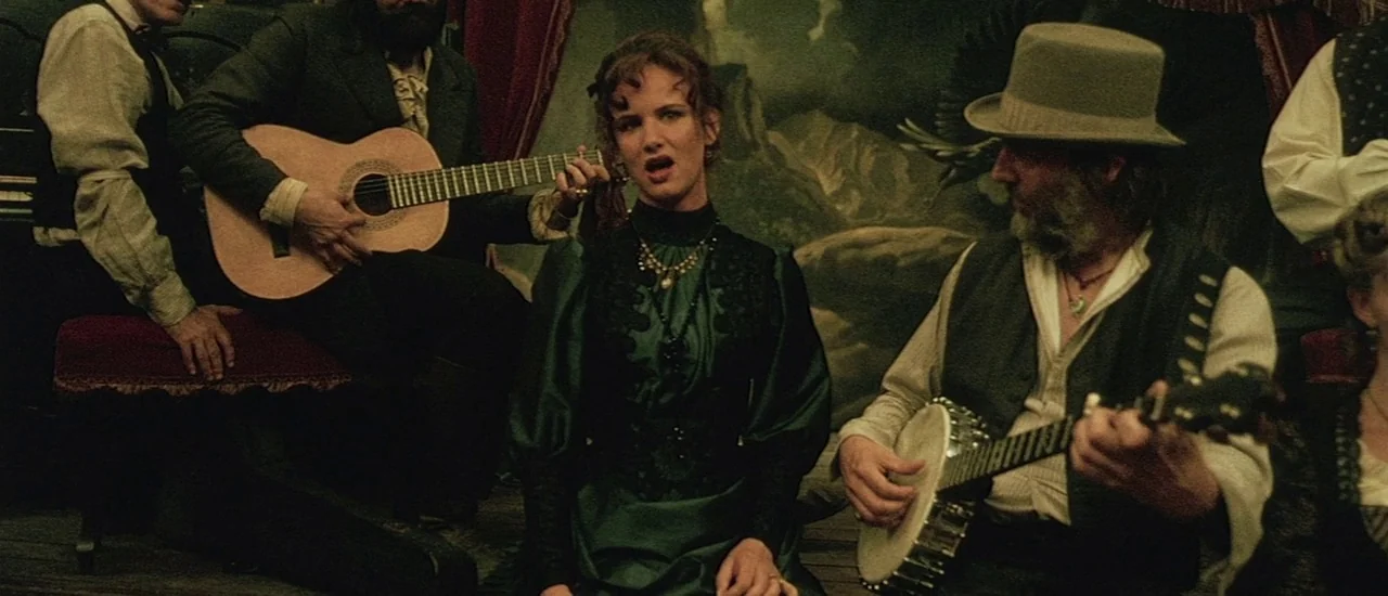 Renegade (2004) movie still: medium — A dimly lit room features a painted landscape backdrop behind a group of musicians. A woman in a green dress…