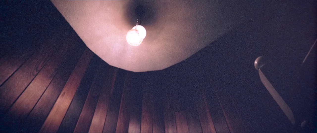 Skinamarink (2022) movie still: establishing — A low angle view of a stairwell ceiling with vertical wood paneled walls. A single glowing pendant…