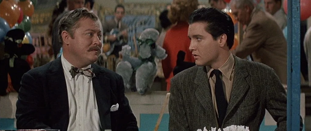 Wild in the Country (1961) movie still: medium — A festive carnival booth with plush poodle prizes decorated in the background. Two men engage in a…