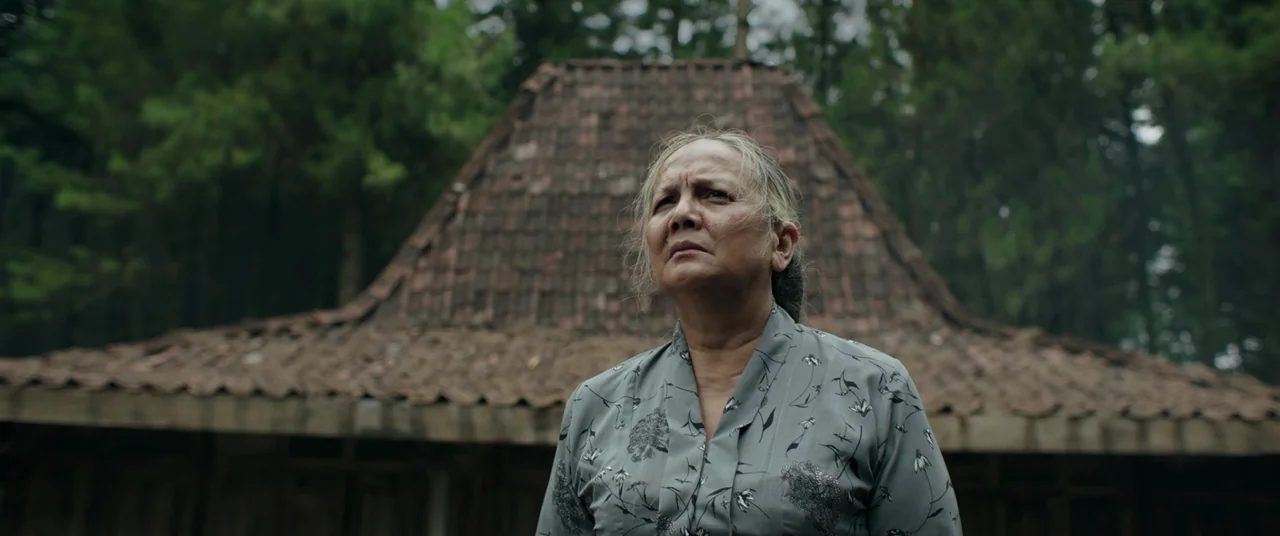 Impetigore (2019) movie still: medium — An elderly woman stands outdoors in front of a rustic wooden structure with tile roofing. The background…