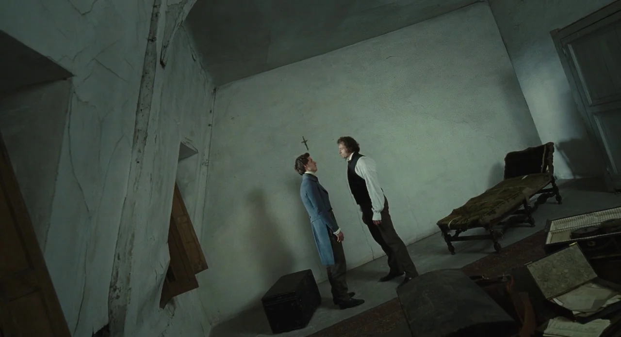 Les Misérables (2012) movie still: fullbody — A sparsely furnished 19th-century room with peeling white plaster walls and a canted floor. Two men…