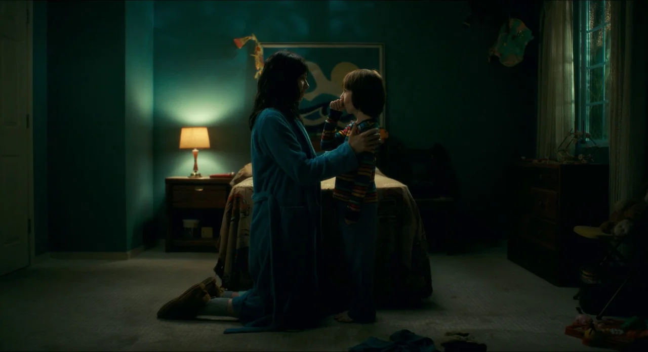 Doctor Sleep movie still: fullbody — A dim room contains a bed with a patterned quilt and a bedside lamp. A woman kneels on the carpet and holds her…