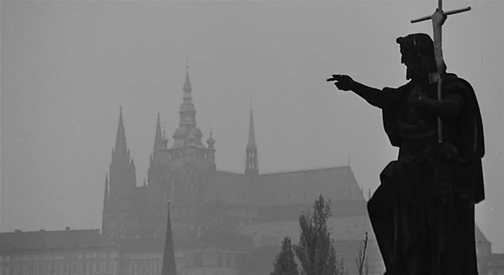 Kafka (1991) movie still: wide — A stone sculpture on a bridge stands in the foreground under grey foggy skies. In the distance a large gothic…