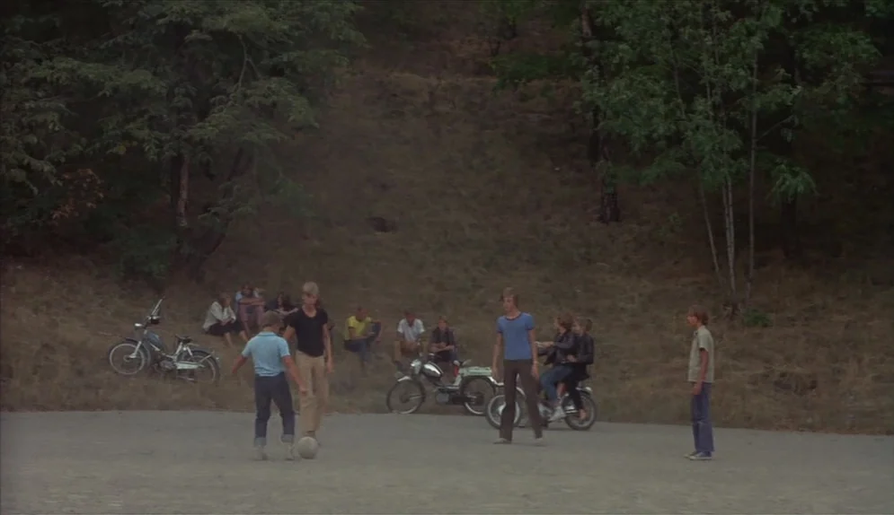 A Swedish Love Story (1970) movie still: wide — A group of teenagers stand on a dirt clearing in front of a wooded hillside. Several motorbikes are…