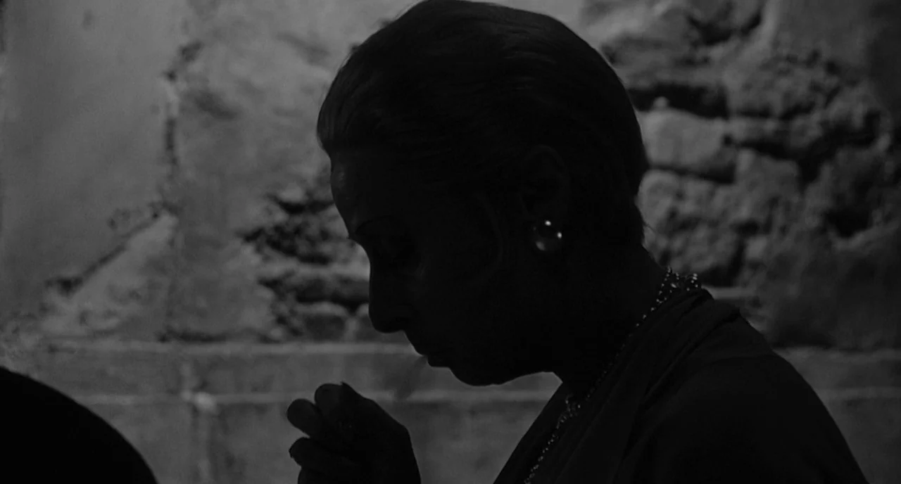 8 1/2 (1963) movie still: closeup — A dimly lit space features a woman in profile against a rough stone wall. The subject keeps her gaze down,…