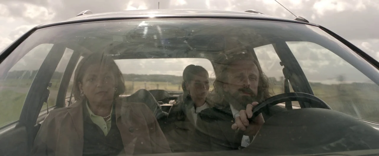 Borgman (2013) movie still: medium — An interior of a car driving through an open rural landscape during the day. Three passengers sit in the front…