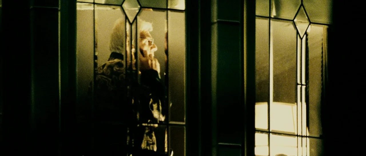 Slice (2018) movie still: medium — An elderly man stands behind a glass-paneled door inside a dimly lit space. He holds a phone to his ear while…