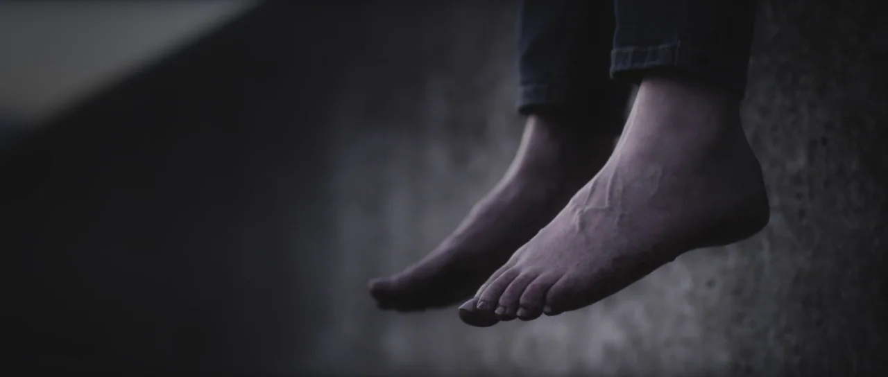 A Dark Song (2016) movie still: closeup — The bare feet of a person hang over the concrete edge of a structure. Dark fabric of jeans covers the…