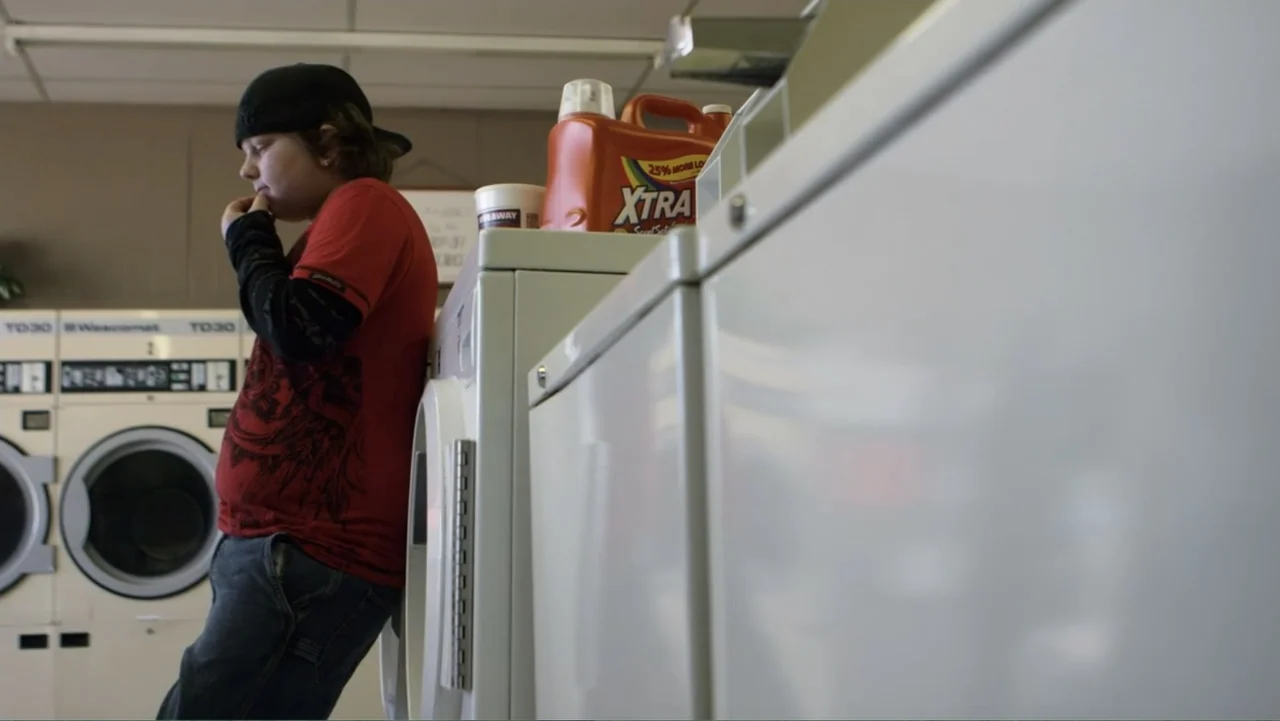Rich Hill (2014) movie still: medium — A utilitarian laundromat interior with rows of white industrial drying machines. A teenage boy leans against…