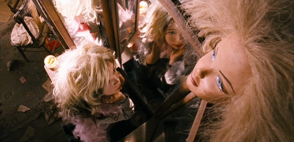 Tideland (2005) movie still: medium — A cluttered interior space features a wooden vanity with a large, upright mirror. A woman with blonde hair…