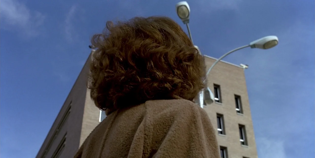 The Flower of My Secret (1995) movie still: closeup — An outdoor urban area with a multi-story brick building under a clear blue sky. A person with…