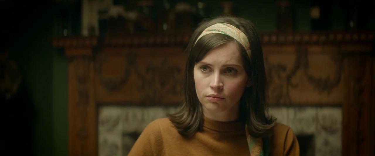 The Theory of Everything (2014) movie still: closeup — A woman stands before a dark green wall and an ornate wooden fireplace mantle. She looks…